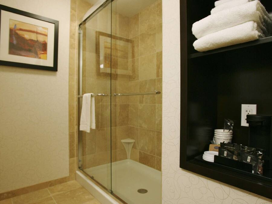 A glass-enclosed shower with a white towel hanging on the door, a framed picture on the wall, and a shelf with towels and toiletries.