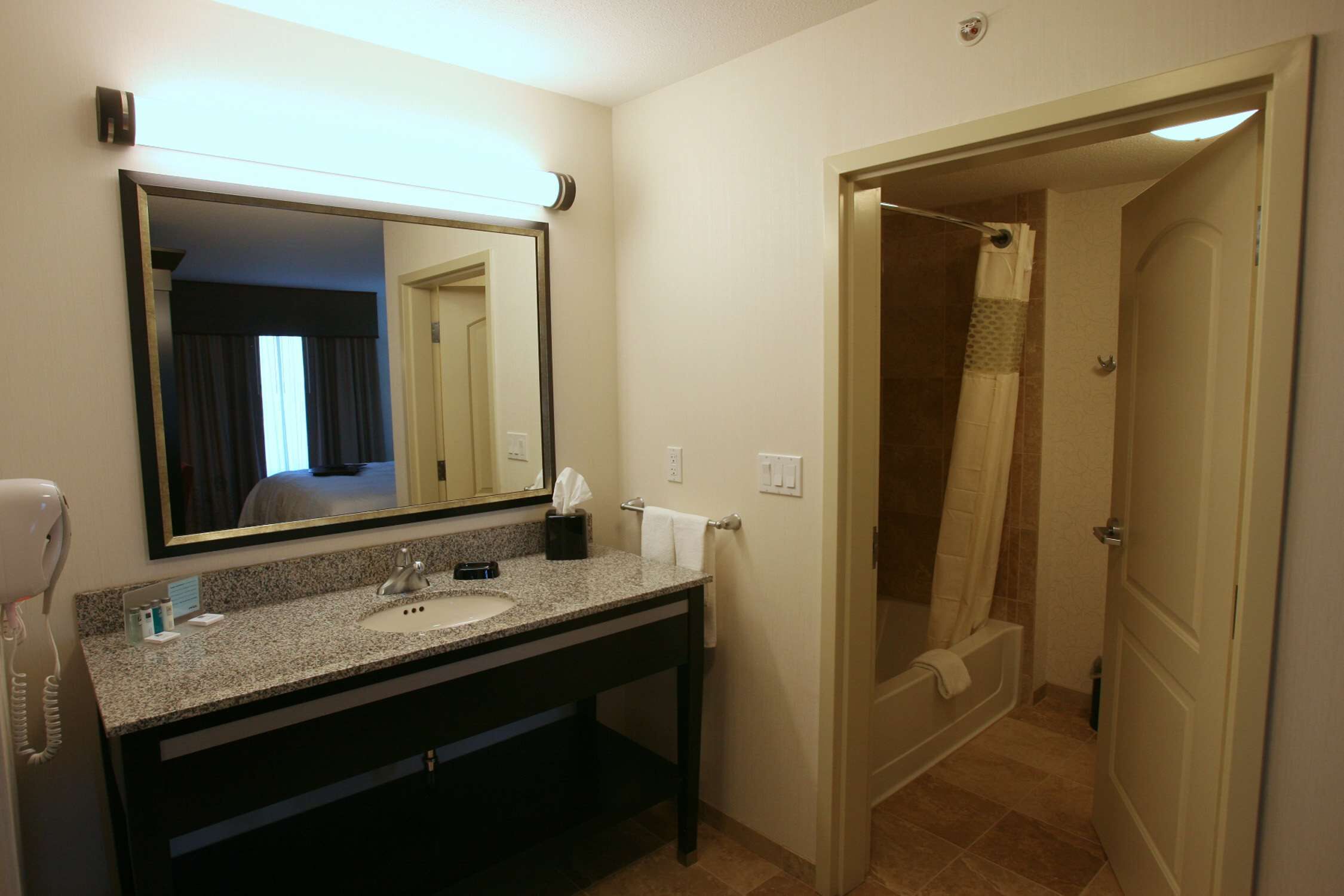 A hotel bathroom with a granite countertop sink, a mirror, a hairdryer, and a shower area.