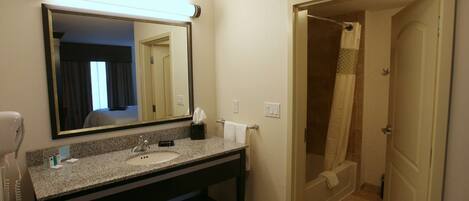 A hotel bathroom with a granite countertop sink, a mirror, a hairdryer, and a shower area.