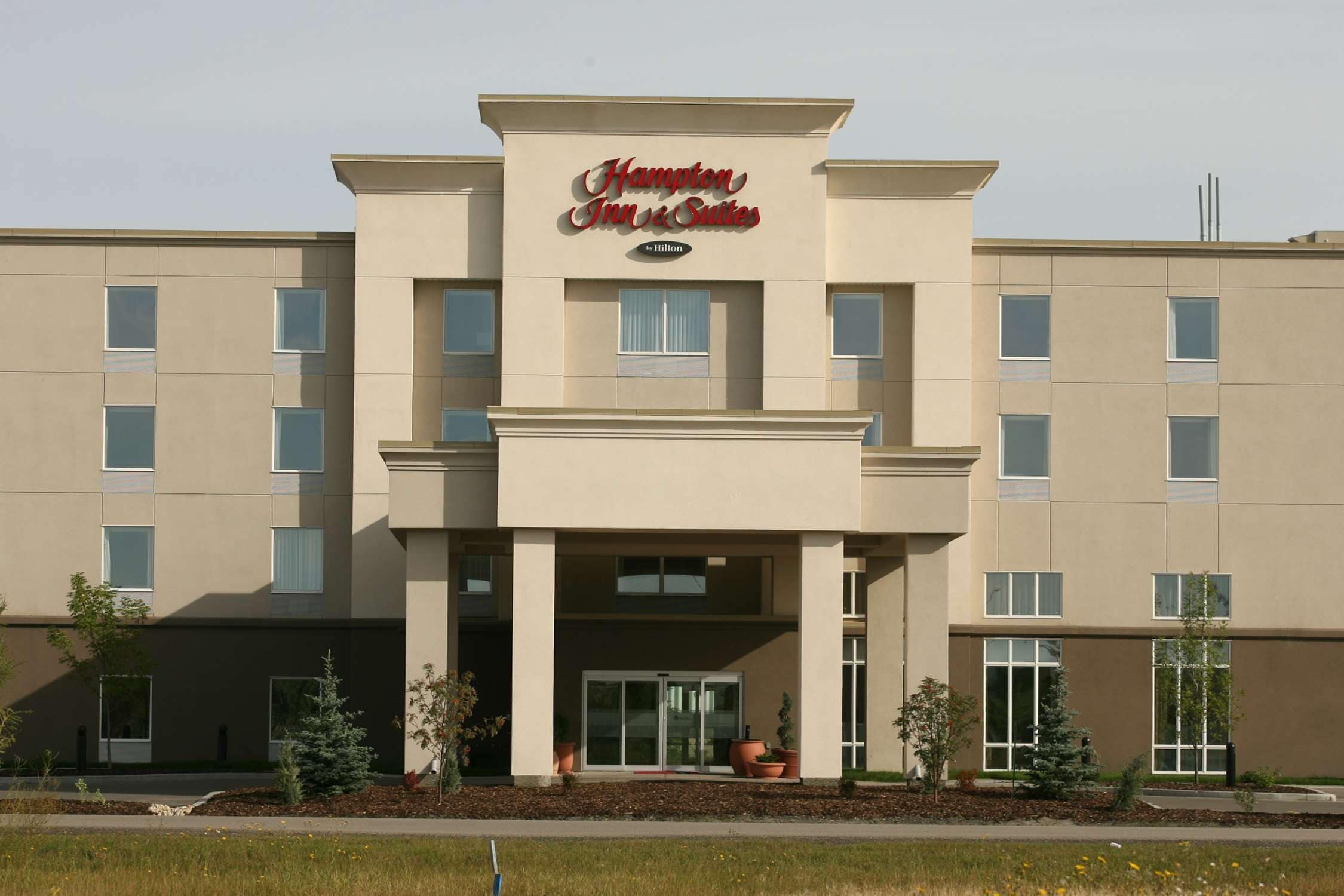 Edificio de Hampton Inn & Suites con fachada color beige, amplias ventanas y una entrada destacada con puerta de vidrio.