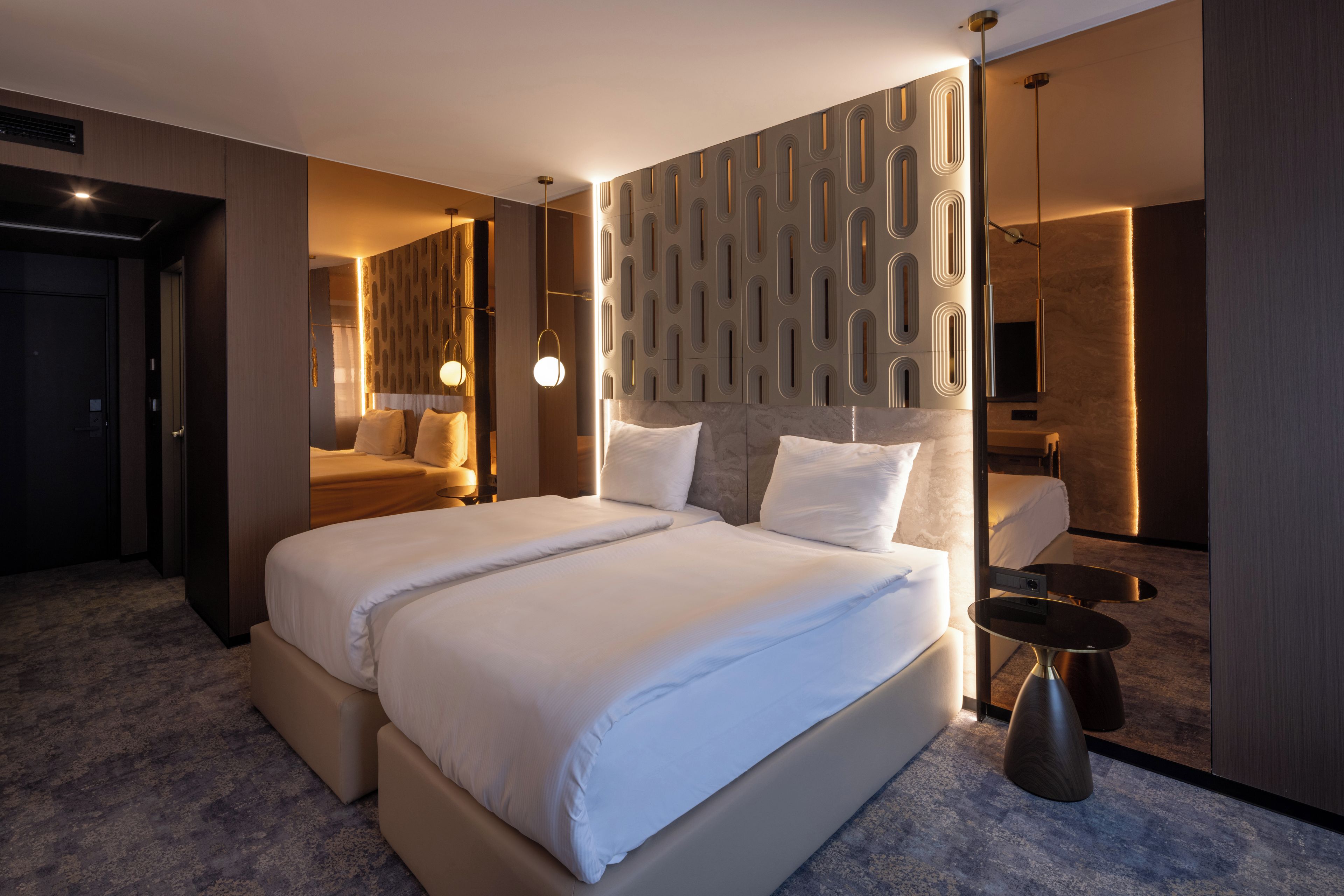 A modern hotel room with a large bed, a bedside table, a mirror, and a wall with a geometric pattern.