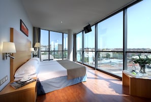 Quarto de hotel moderno com uma cama grande, duas luminárias de cabeceira, uma televisão de tela plana e vista da cidade através de amplas janelas.