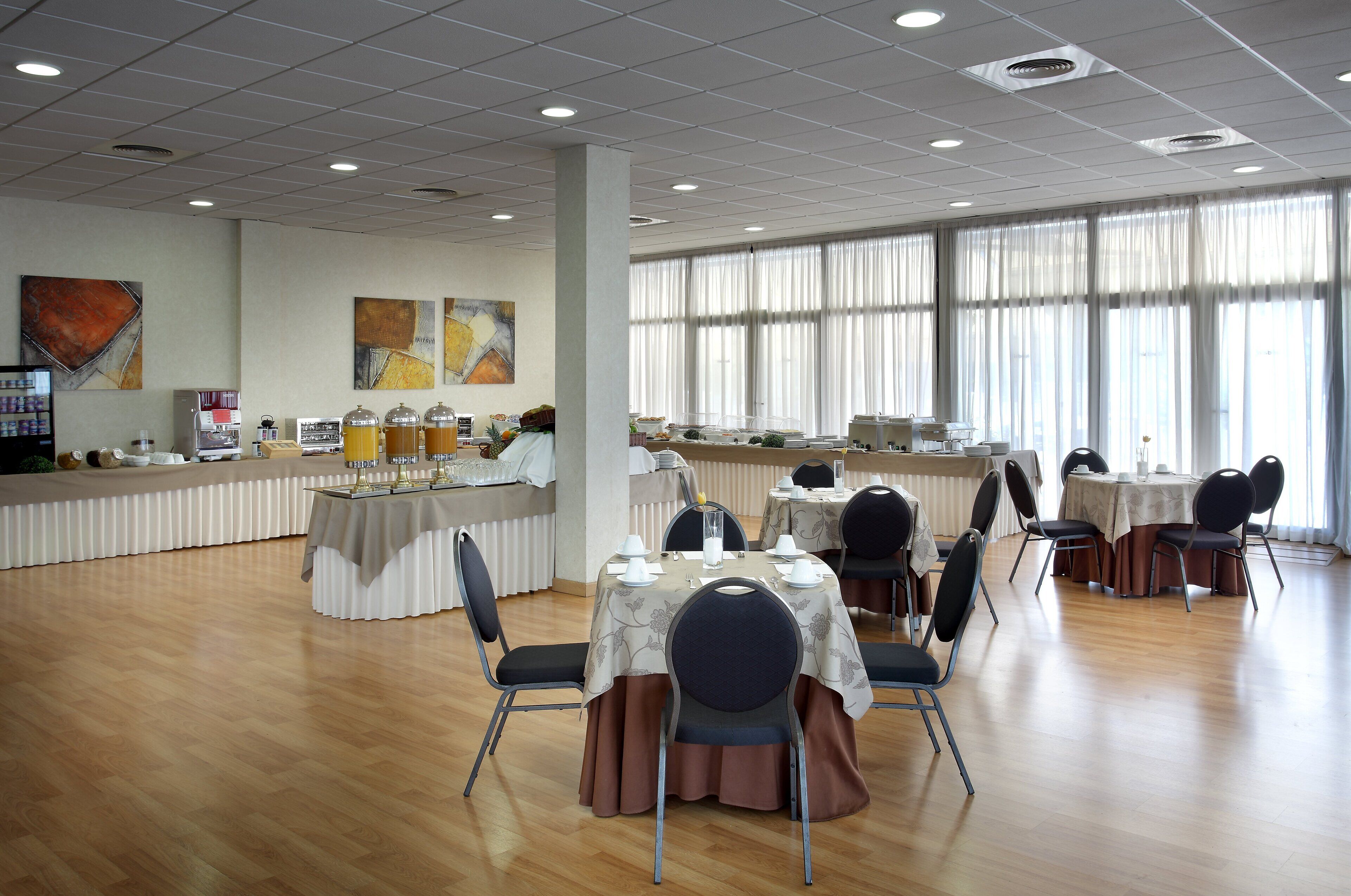 Un'ampia sala da pranzo con diversi tavoli e sedie, grandi finestre con tende leggere e un bancone con distributori di cibo e bevande.