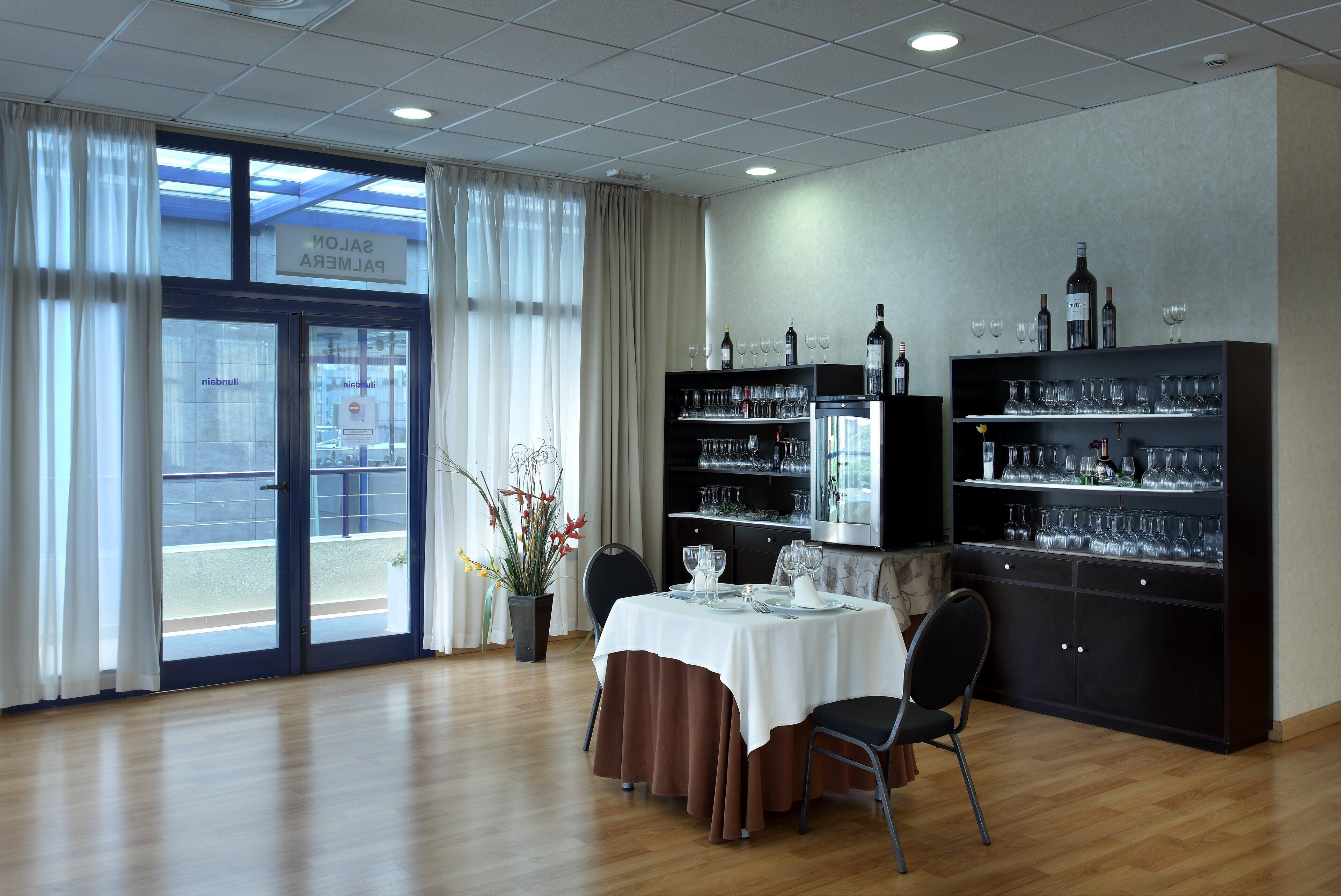 Un coin repas avec une table dressée pour deux personnes, une plante en pot et une vue sur l’extérieur, visible à travers une porte vitrée.