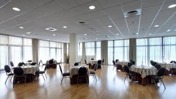 Un amplio salón de conferencias con numerosas mesas y sillas, ventanas grandes y un techo alto con iluminación empotrada.