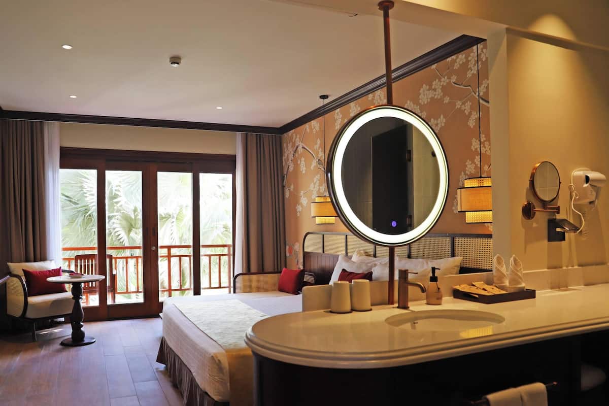 A hotel room with a large bed, a round mirror, a vanity with a sink, and a balcony with curtains.