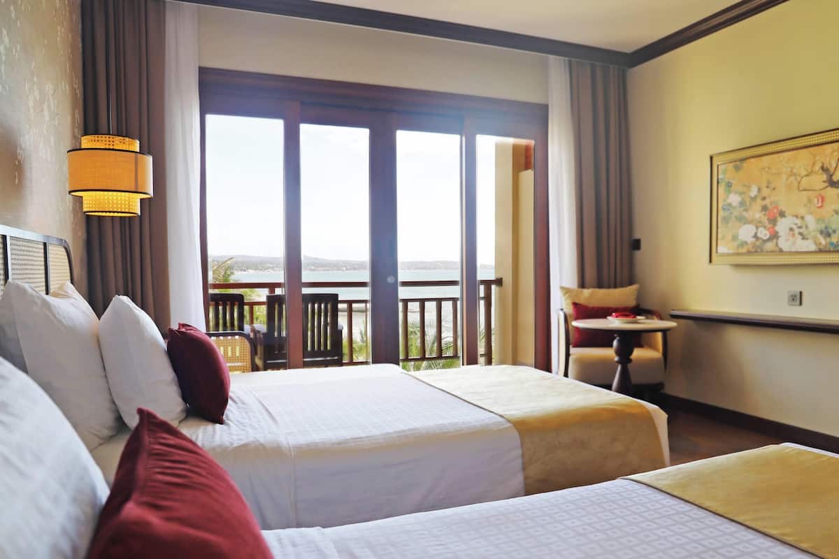 A hotel room with two beds, a balcony with a view, a small table, and a painting on the wall.