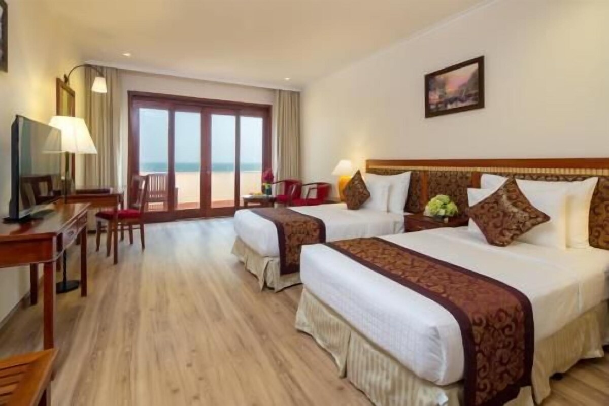Habitación de hotel con dos camas, un escritorio, una silla, un televisor y un ventanal con vista a la playa.