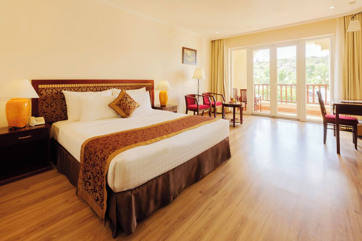 A hotel room with a large bed, a desk with a chair, a small table with a chair, and a balcony with a view of greenery.