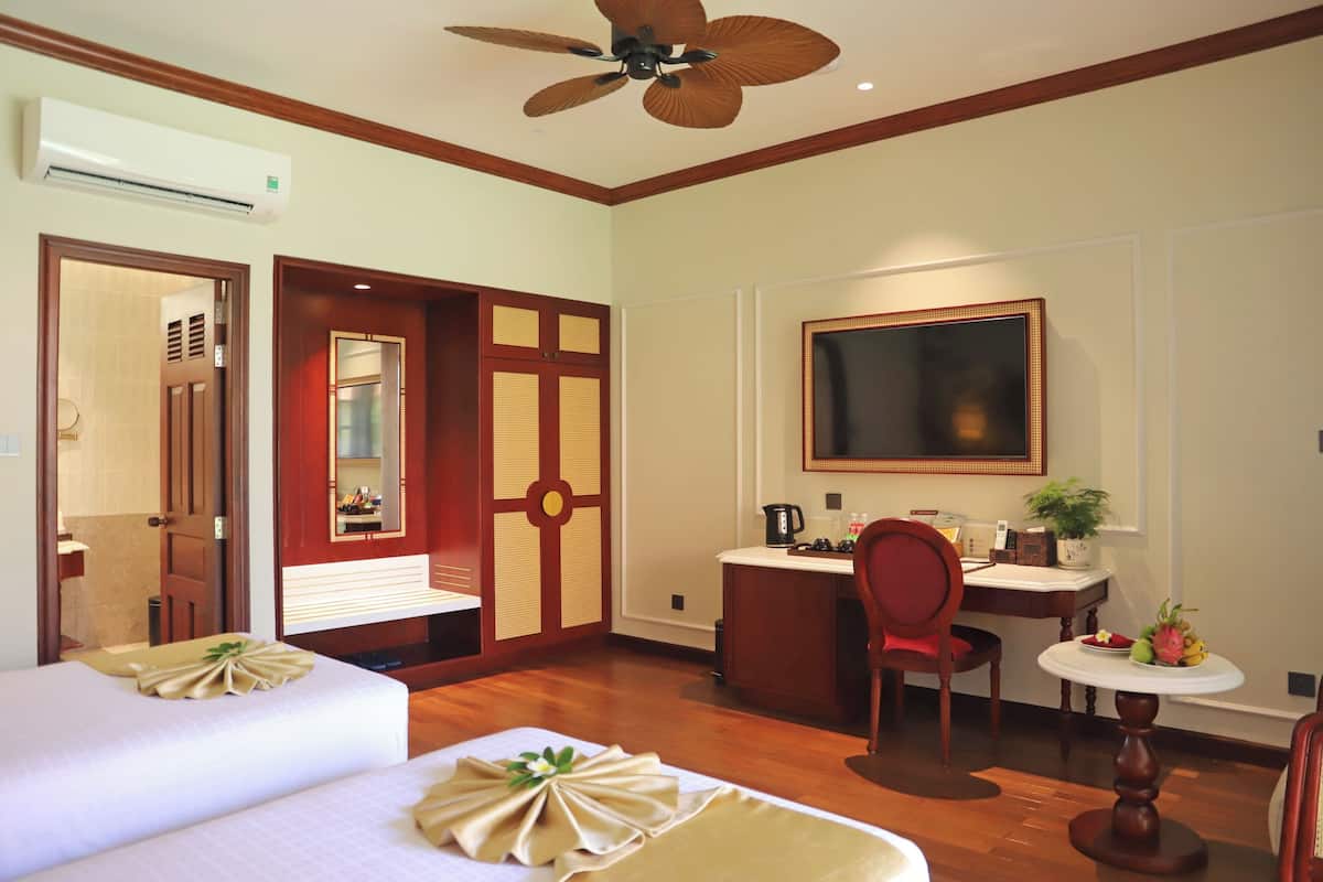 A hotel room with two beds, a desk, a chair, a ceiling fan, a TV, and a small table with fruit.