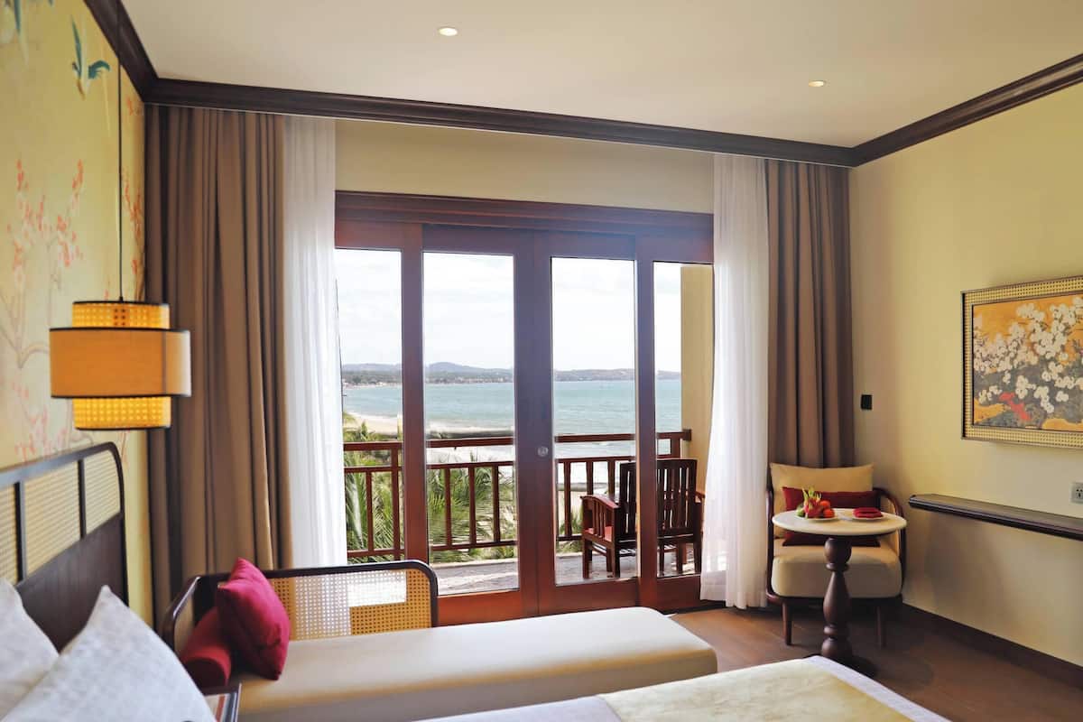 A hotel room with two beds, a balcony with a view of the sea, a dining table, and a painting on the wall.