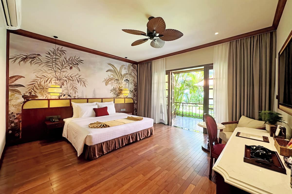 A hotel room with a bed, bedside table, lamp, ceiling fan, and a view of the outdoors.