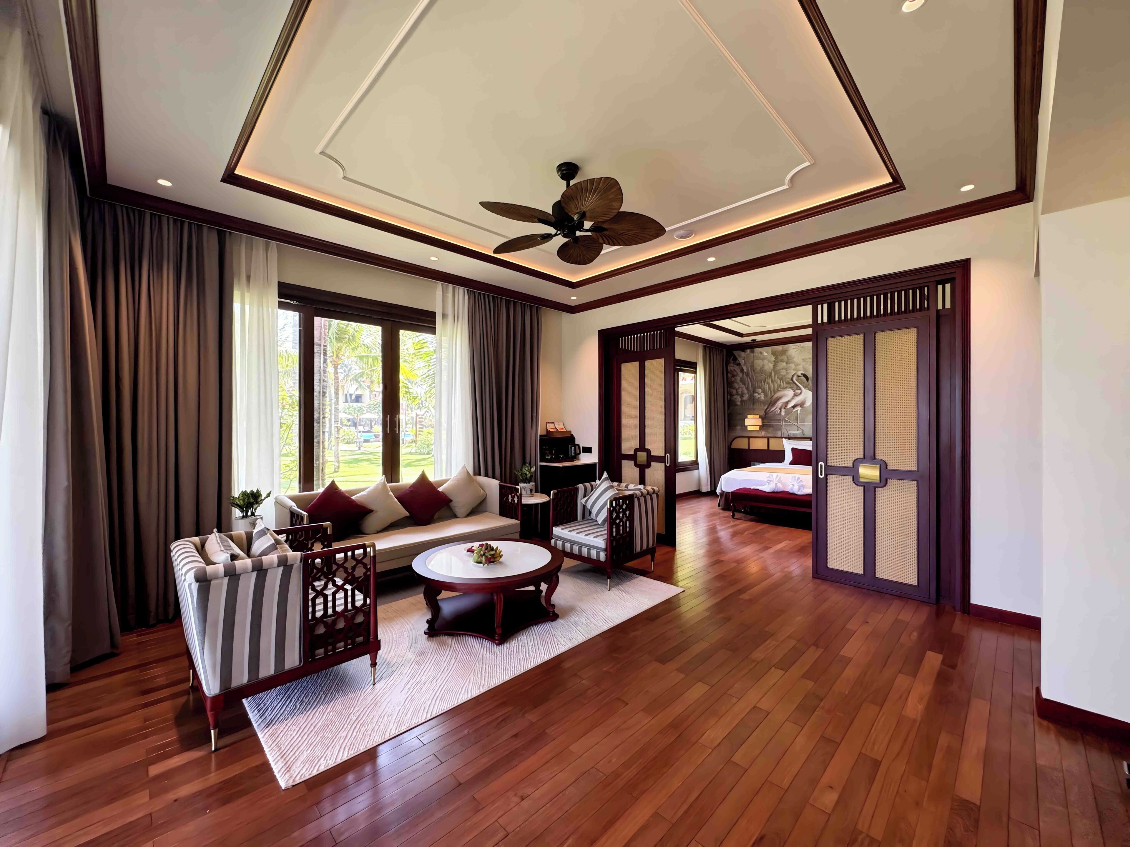 A spacious hotel room with a large bed, a sofa, a dining table, and a ceiling fan.