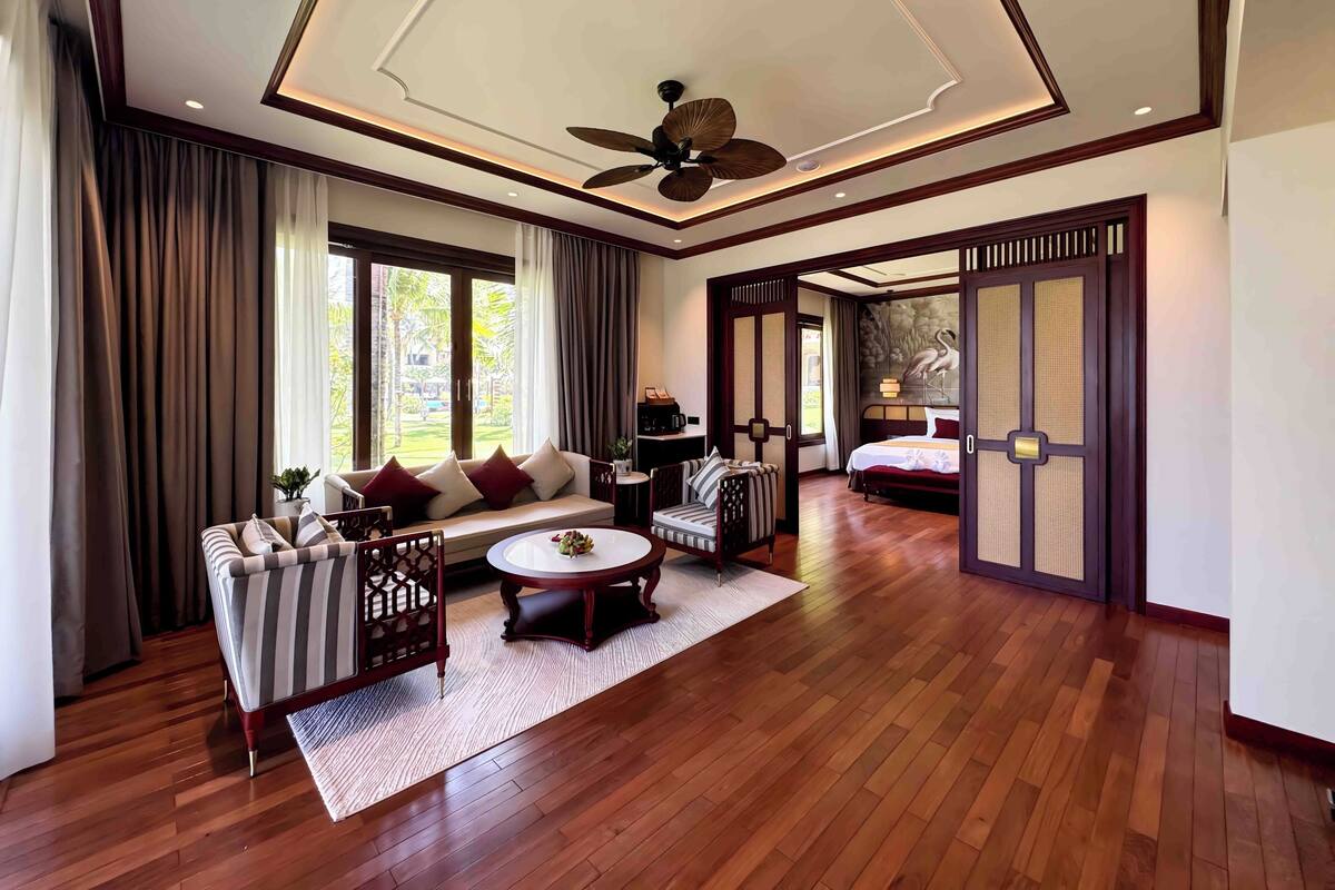 A spacious hotel room with a large bed, a sofa, a dining table, and a ceiling fan.