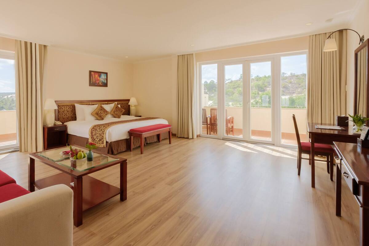 A hotel room with a large bed, a desk, a chair, a balcony with a table and chairs, and a view of the outdoors.