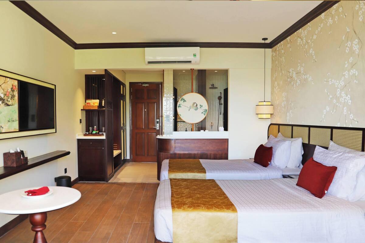 A hotel room with two beds, a round table, a mirror, and a bathroom visible through an open door.