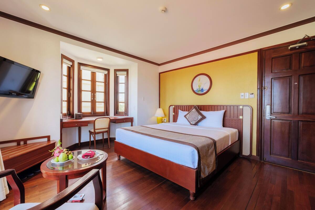 A hotel room with a bed, desk, chair, TV, and a dining table with fruit.