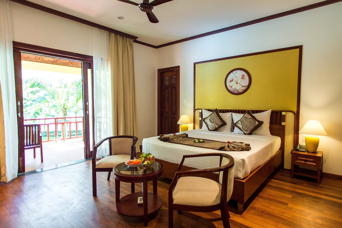 A hotel room with a bed, two chairs, a small table, a bedside lamp, a clock, and a view of the outdoors.
