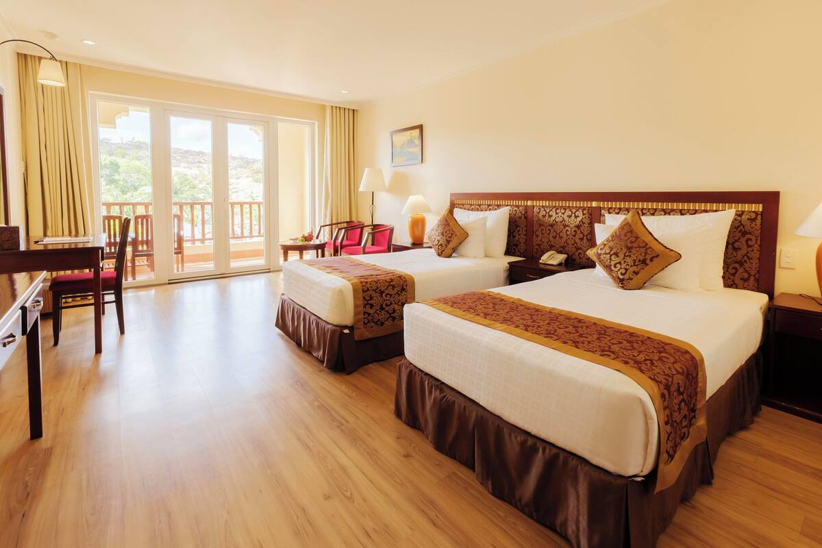 A hotel room with two beds, a desk, chairs, a balcony, and a view of the outdoors.