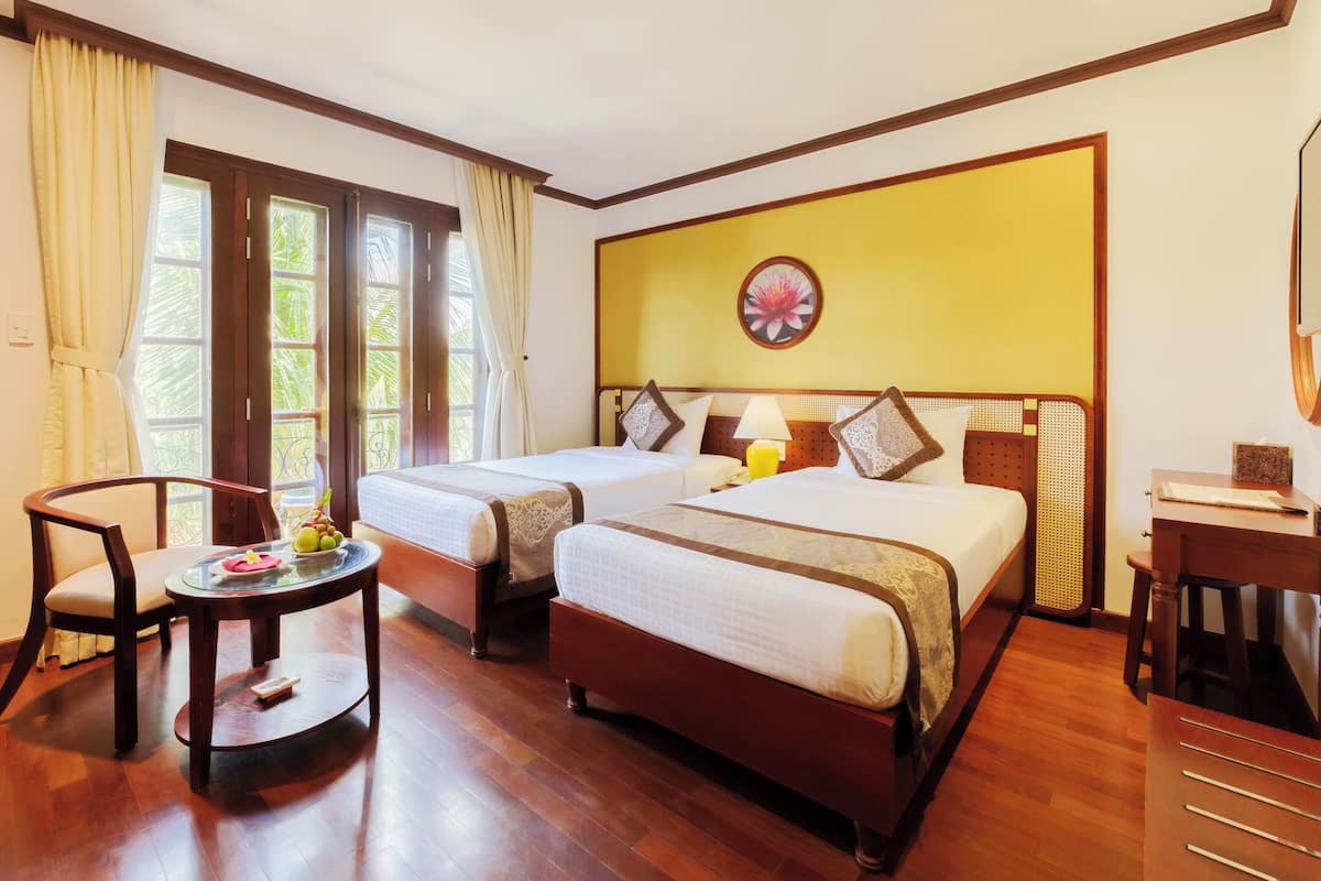 A hotel room with two beds, a small table with fruit, a chair, a desk, and a window with curtains.