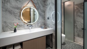 Un baño moderno con pared de mármol, un espejo redondo con luz, un lavabo blanco y una cabina de ducha de vidrio.