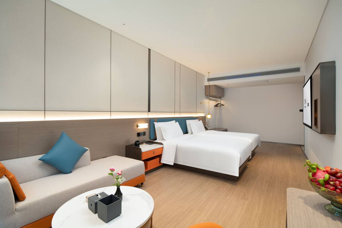 Ein modernes Hotelzimmer mit einer Couch, zwei Betten, einem Nachttisch, einem kleinen Tisch mit einer Vase und einer Obstschale.