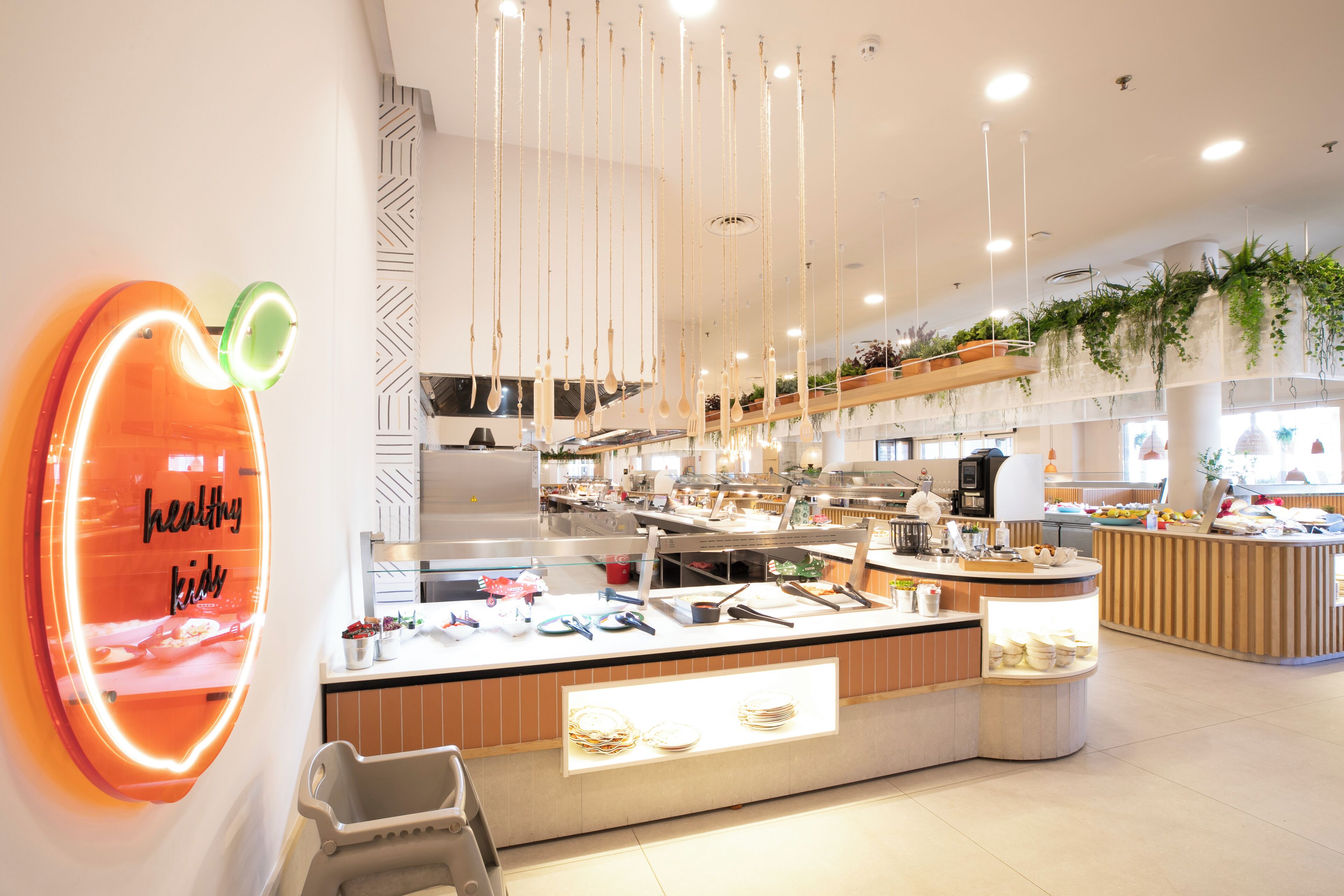 A modern cafe with a neon sign reading "healthy kids," a variety of food items on display, and hanging plants.