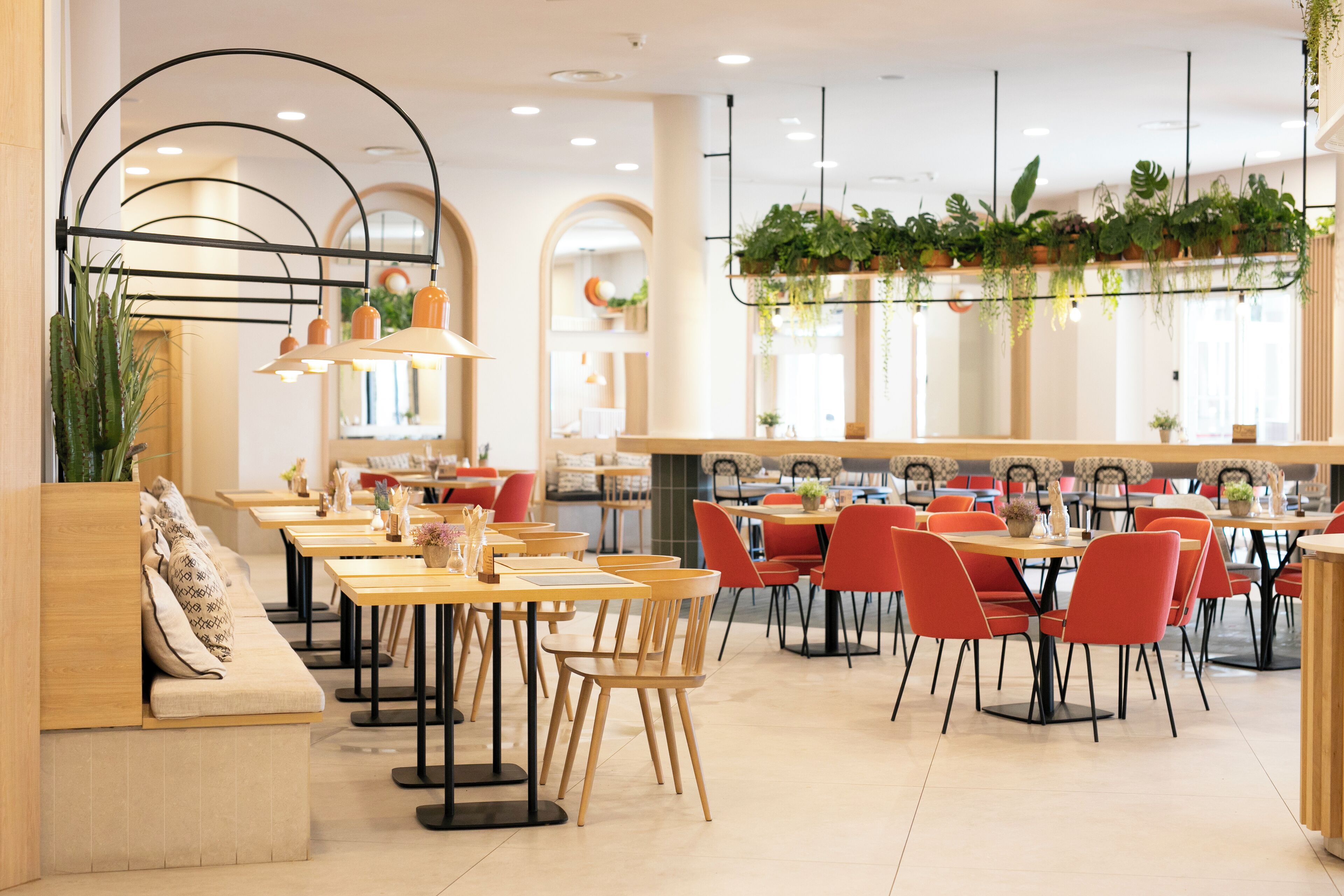 Een modern restaurant met houten tafels en rode stoelen, met een lange toonbank met een bankje en hangplanten.