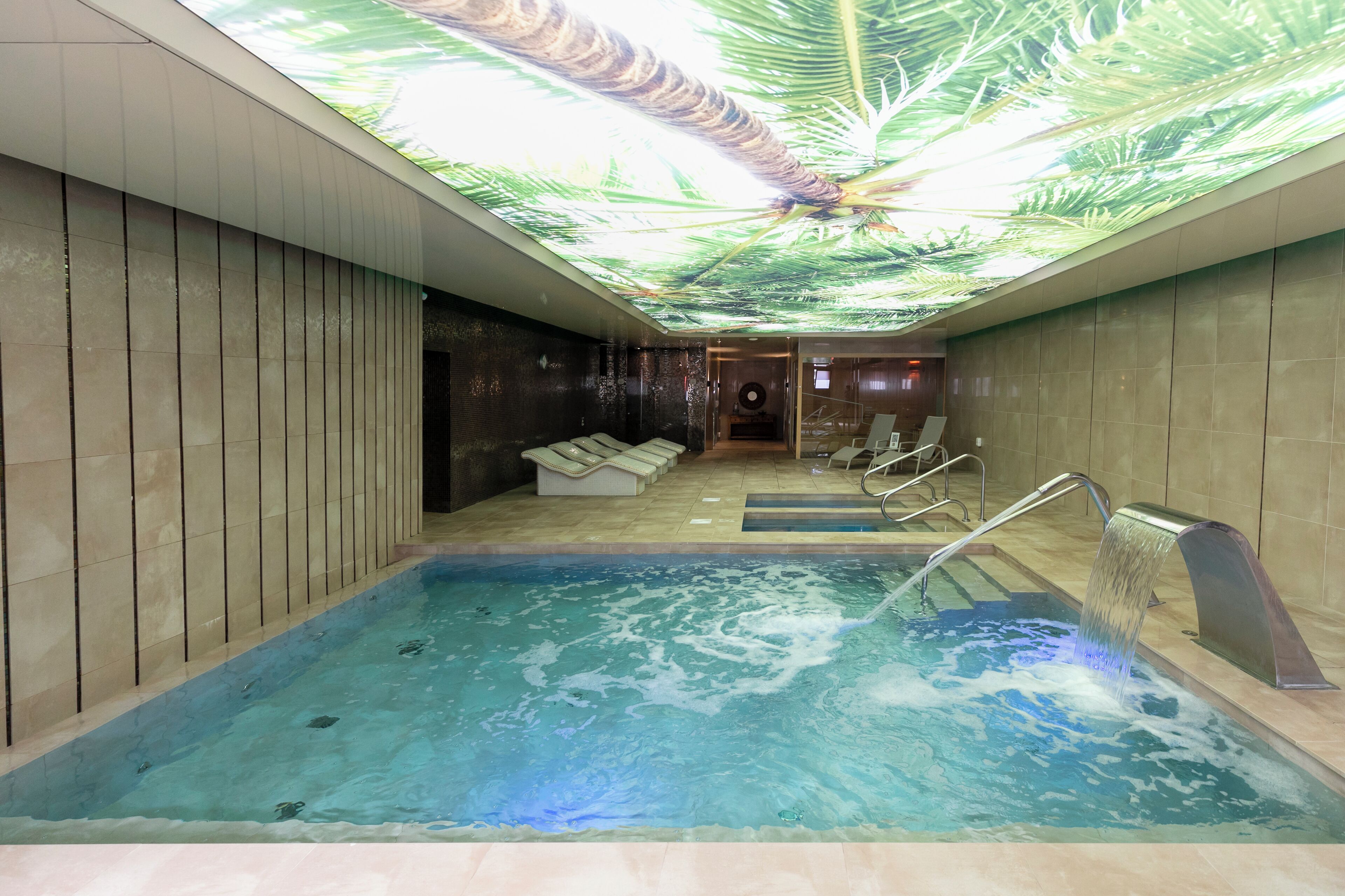 A modern indoor pool with a ceiling mural, a seating area, and a water feature.