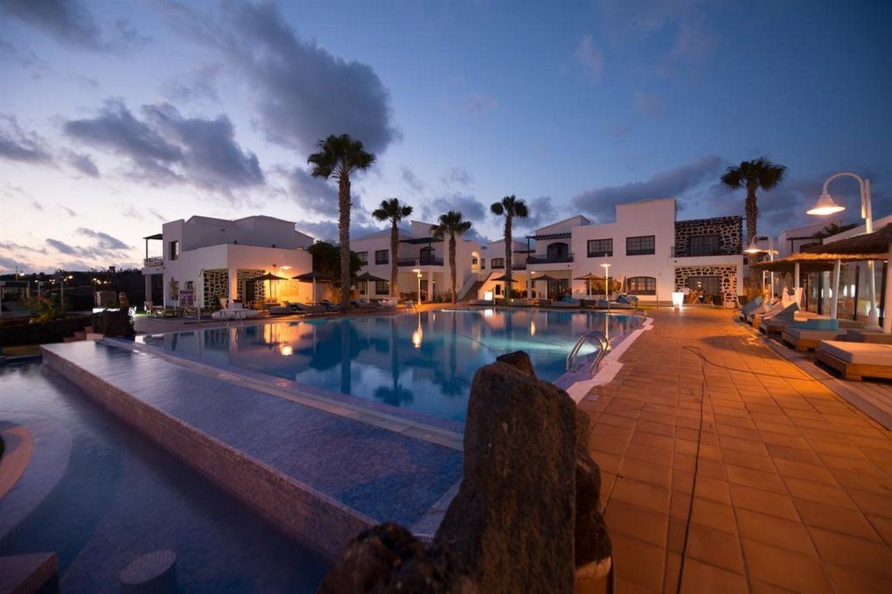 Een modern hotel met een groot zwembad, palmbomen en een zonsondergang.