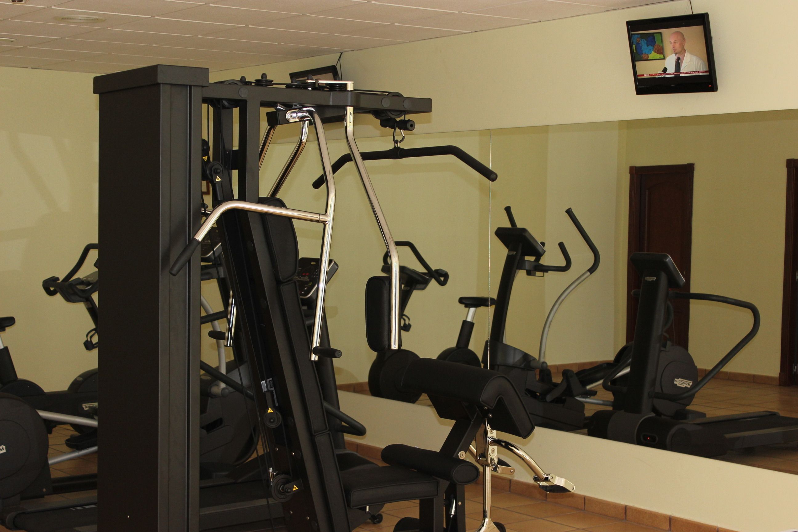 A gym with various exercise machines, a television mounted on the wall, and a large mirror reflecting the equipment.