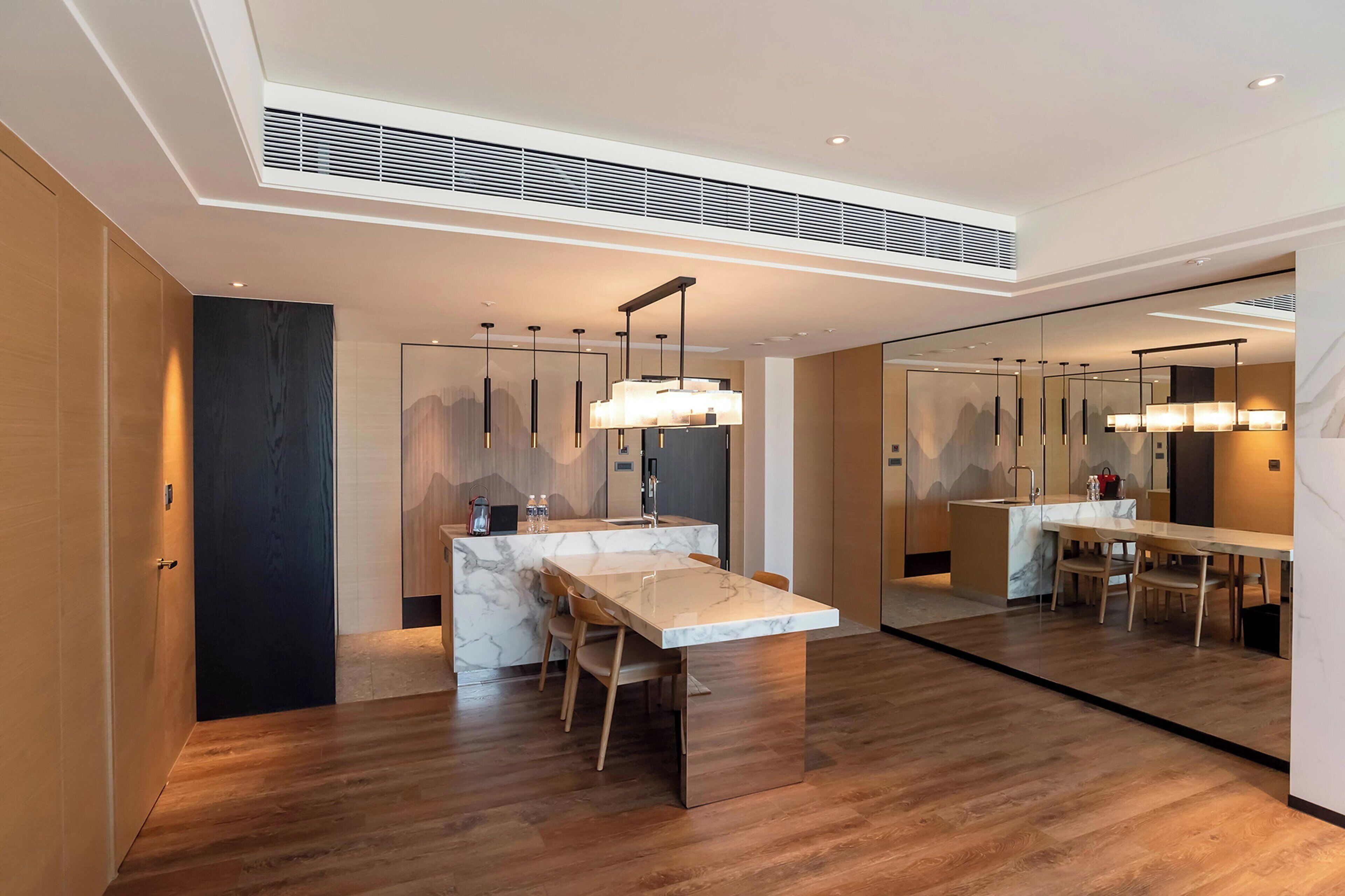 A modern kitchen with a marble countertop, wooden chairs, pendant lighting, and a large mirror reflecting the space.