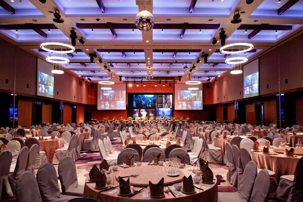 Un amplio salón de banquetes con mesas redondas, sillas y un escenario con una pantalla grande.