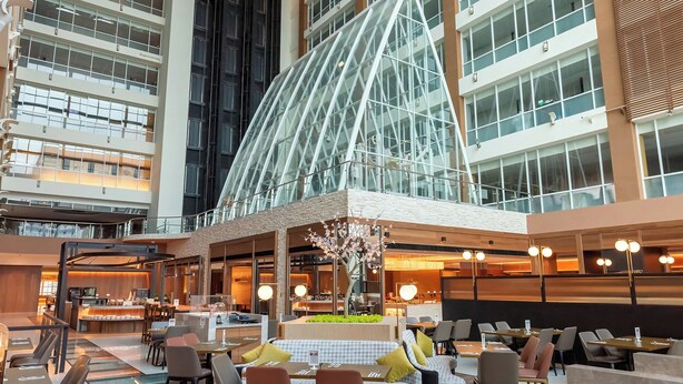 A modern hotel lobby with a glass atrium, seating areas, and a central decorative tree.