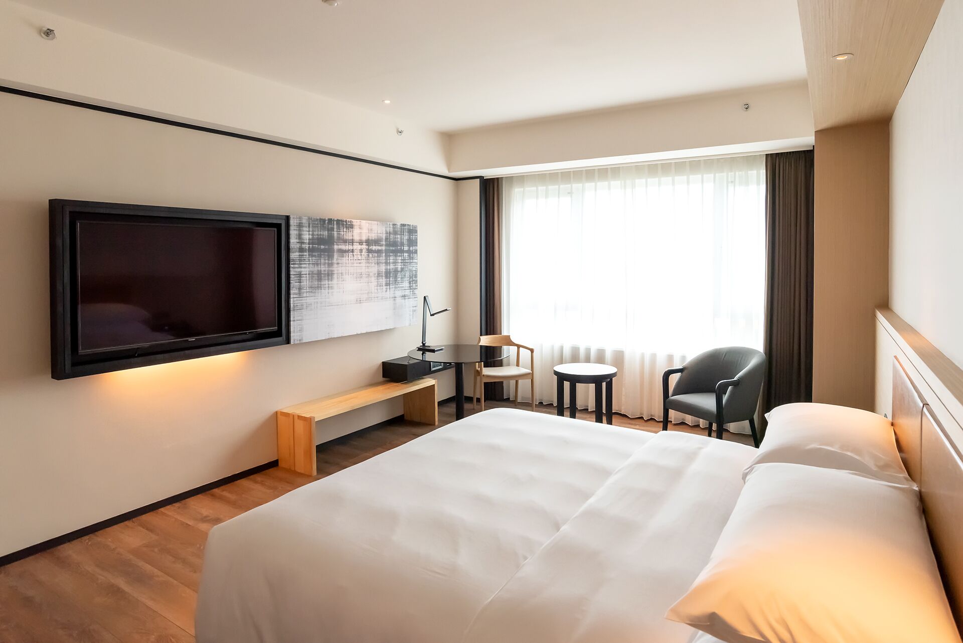 Habitación de hotel con una cama grande, un televisor de pantalla plana montado en la pared, un escritorio pequeño con una silla, una mesa redonda con una silla y un ventanal con cortinas.