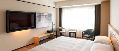 Habitación de hotel con una cama grande, un televisor de pantalla plana montado en la pared, un escritorio pequeño con una silla, una mesa redonda con una silla y un ventanal con cortinas.