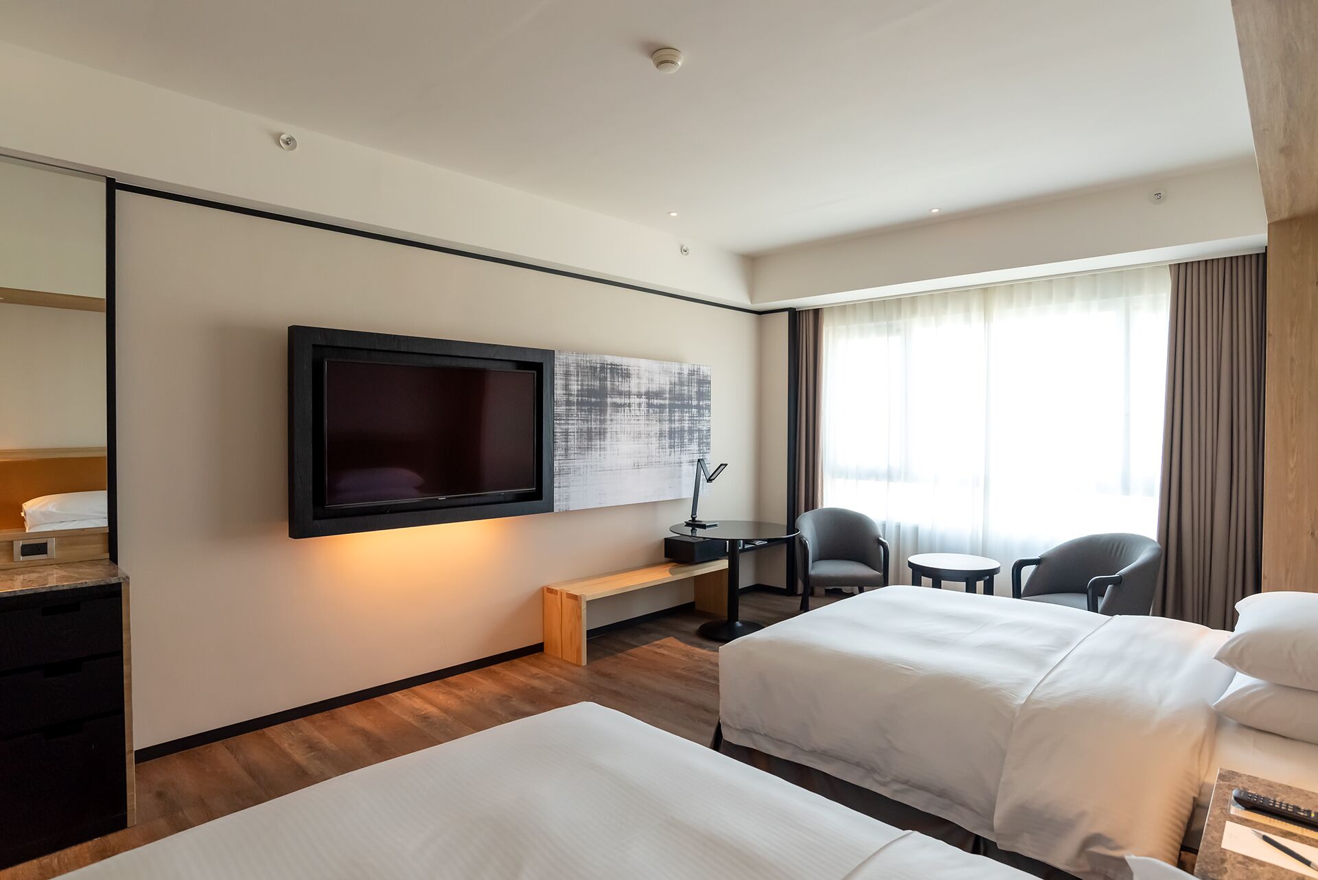 A hotel room with two beds, a TV mounted on the wall, a desk with a chair, and a window with curtains.