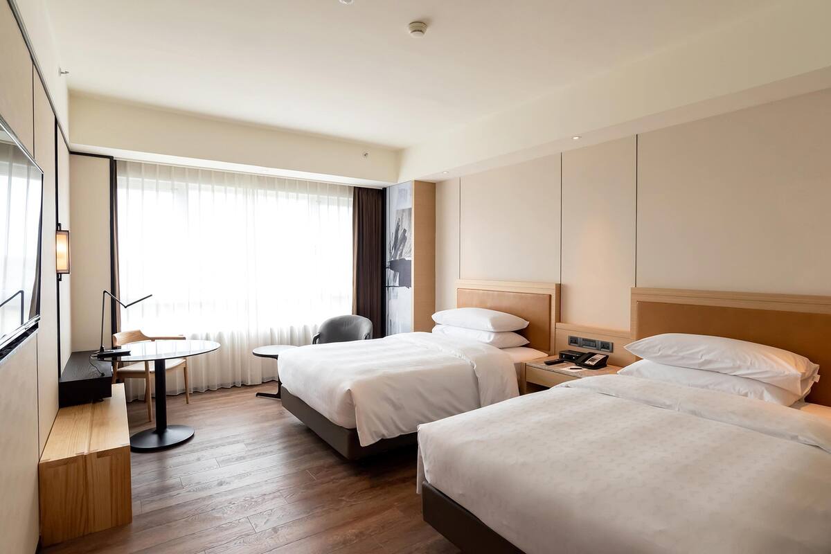 Ein Hotelzimmer mit zwei Betten, einem Schreibtisch, einem Stuhl und einem großen Fenster mit Vorhängen.