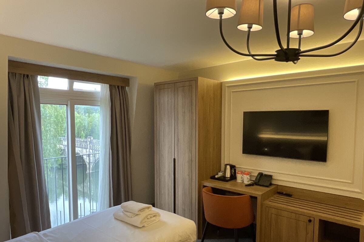 A hotel room with a bed, a desk with a chair, a television mounted on the wall, and a window with curtains.