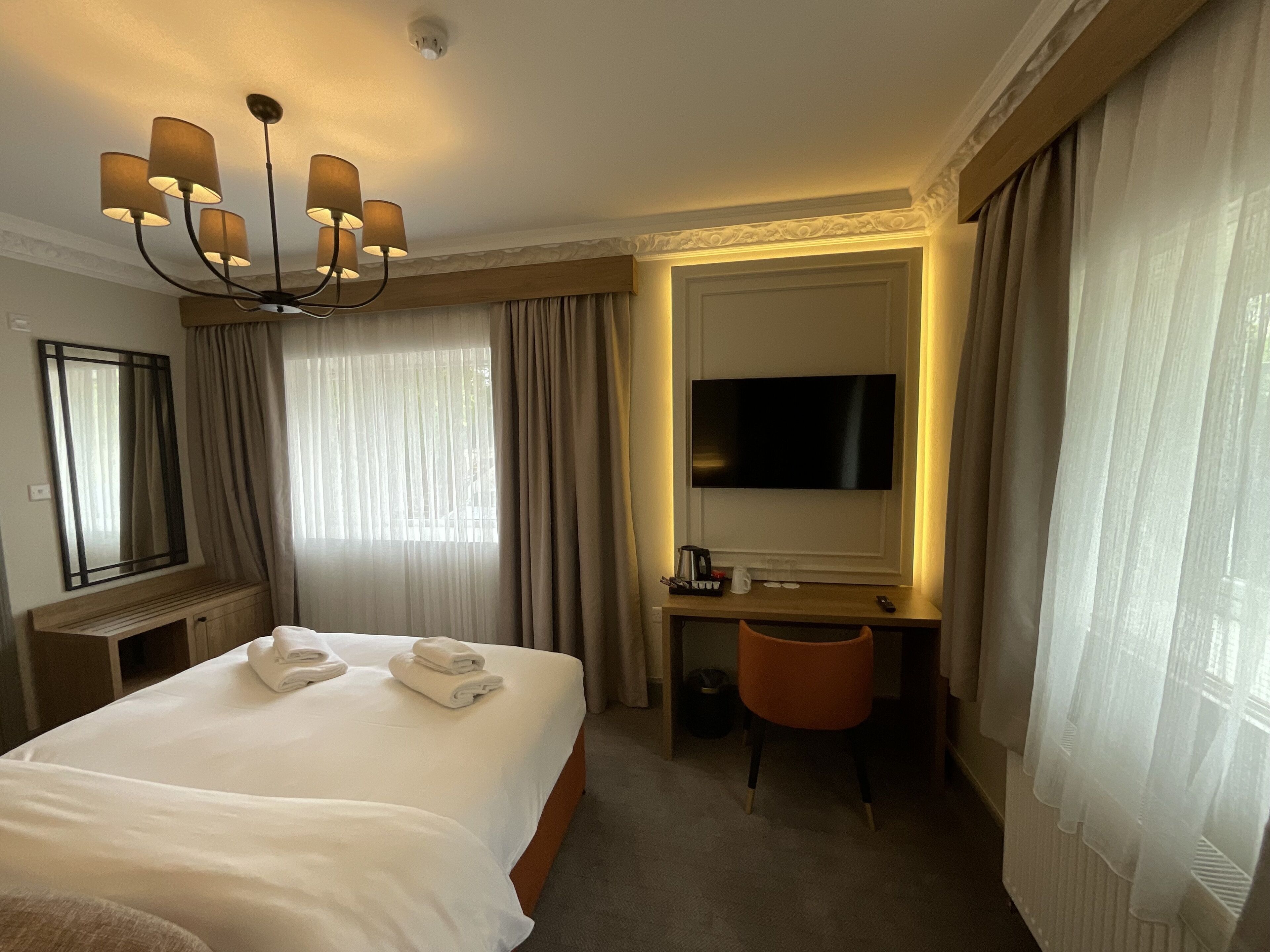 A hotel room with a large bed, a desk with a chair, a television, and a chandelier.