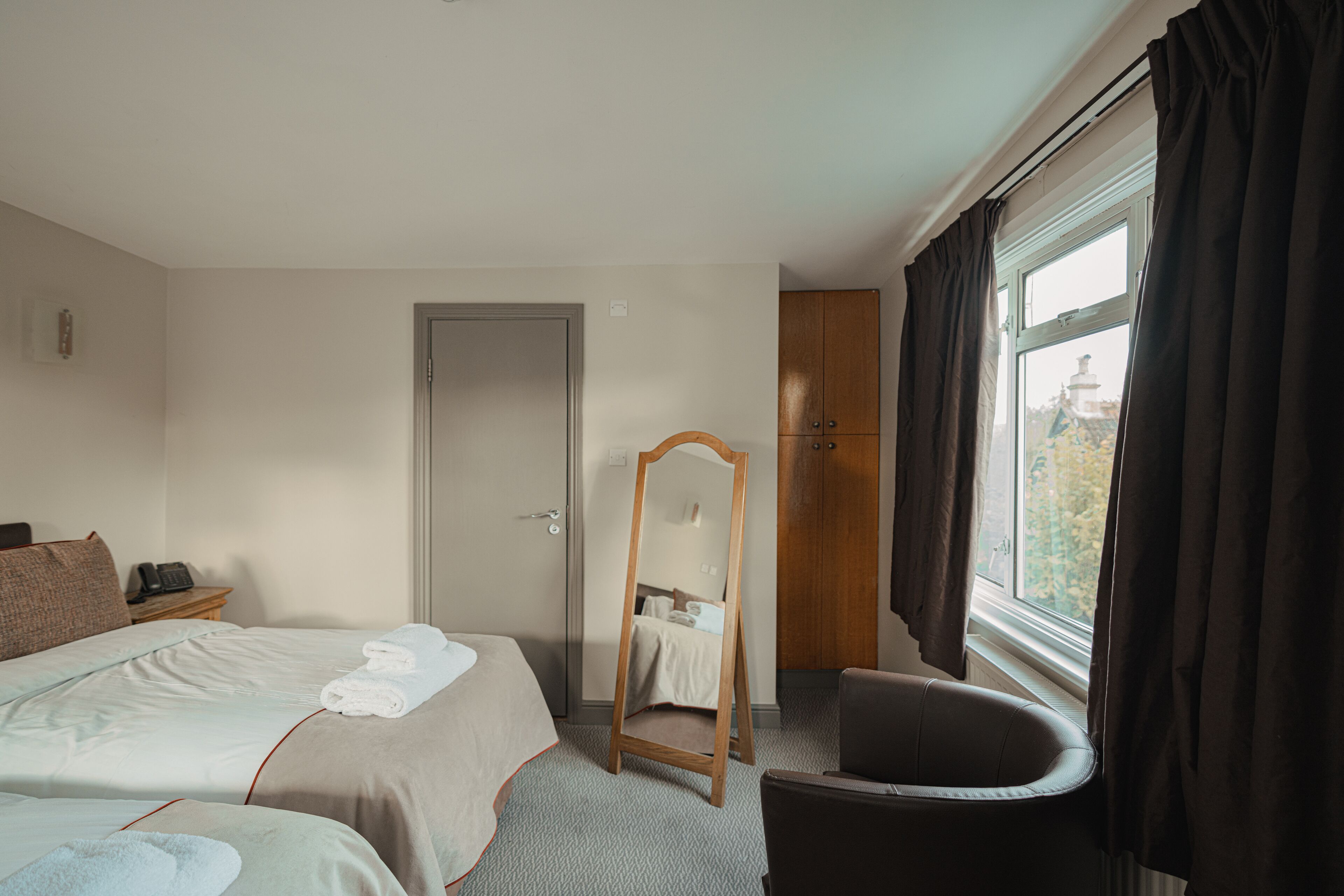 A hotel room with two beds, a wooden chair, a mirror, and a window with curtains.