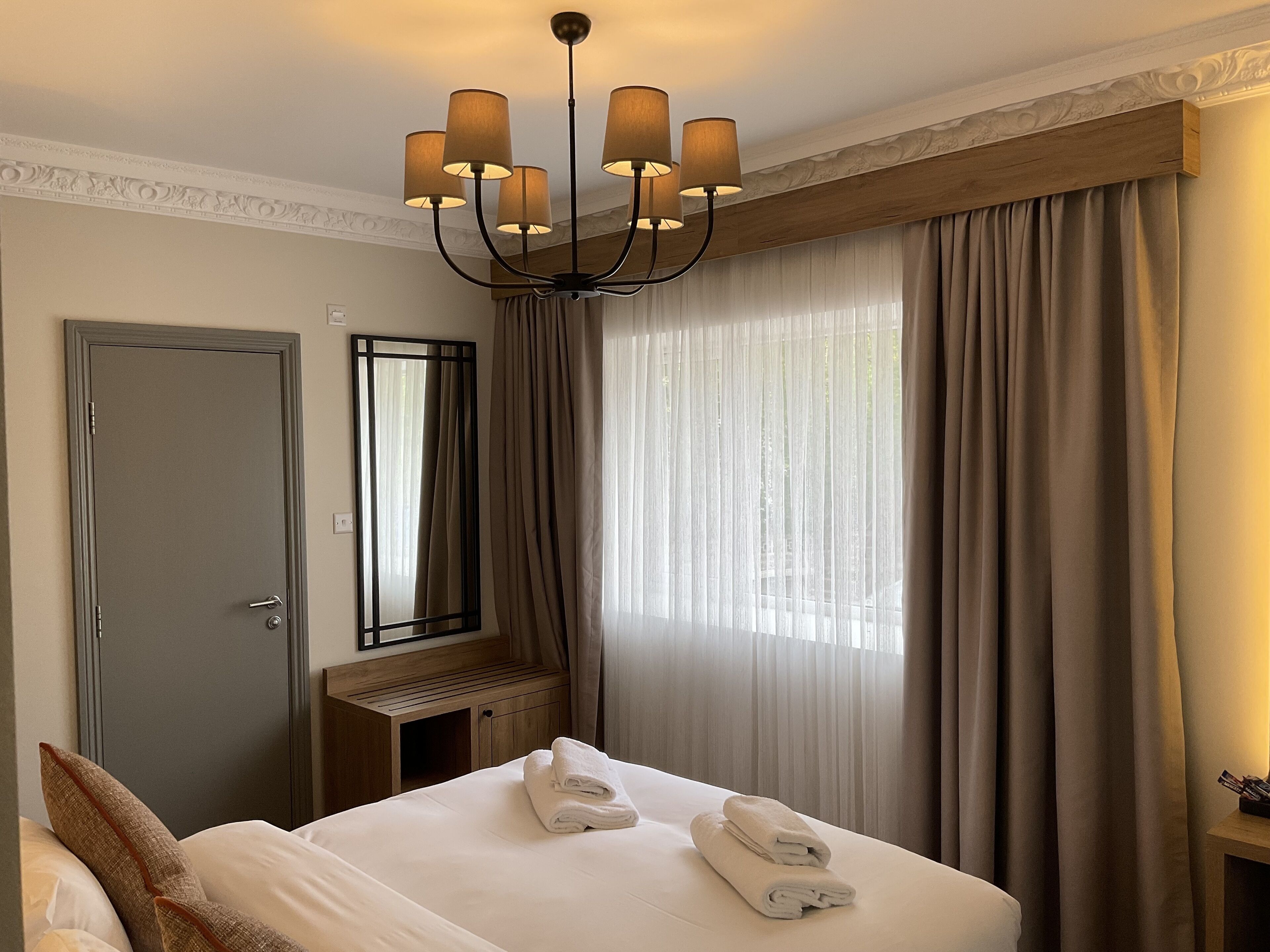 A hotel room with a bed, a mirror, a chandelier, and a window with curtains.