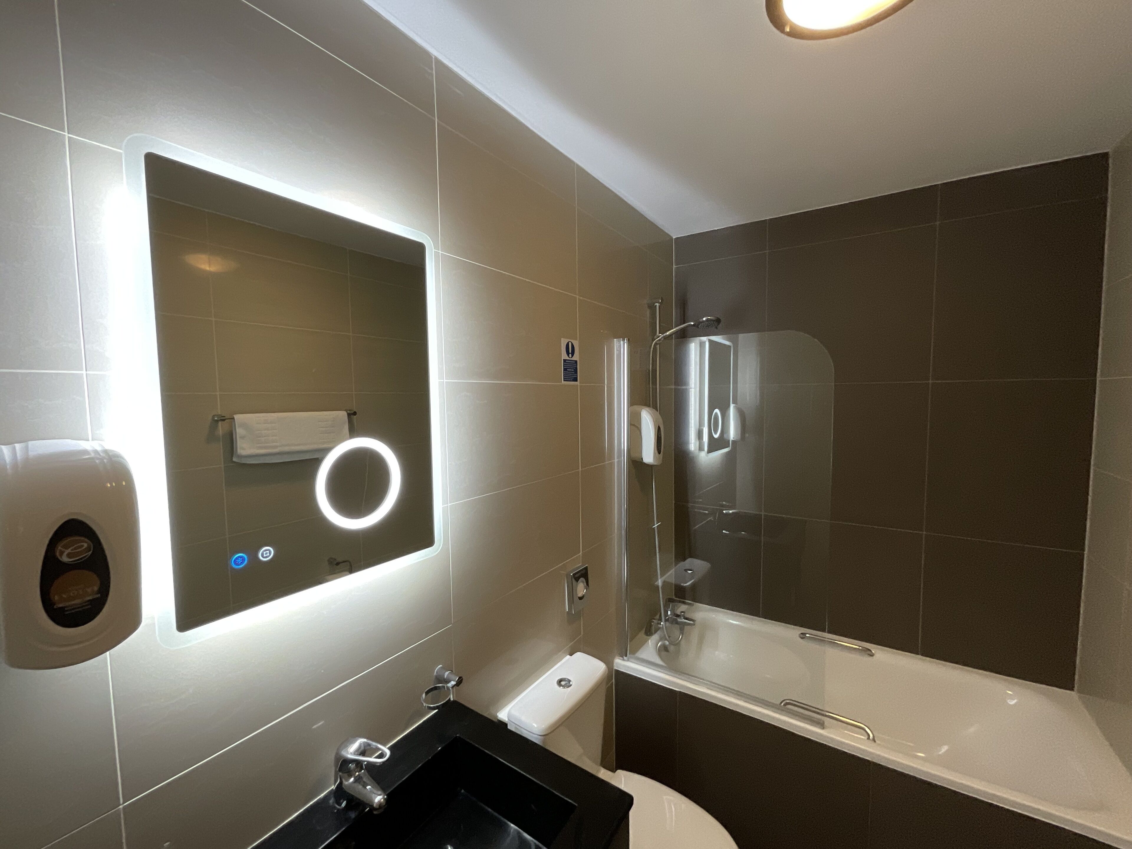 Un baño moderno con ducha acristalada, tocador con encimera negra y espejo con luz integrado en la pared.
