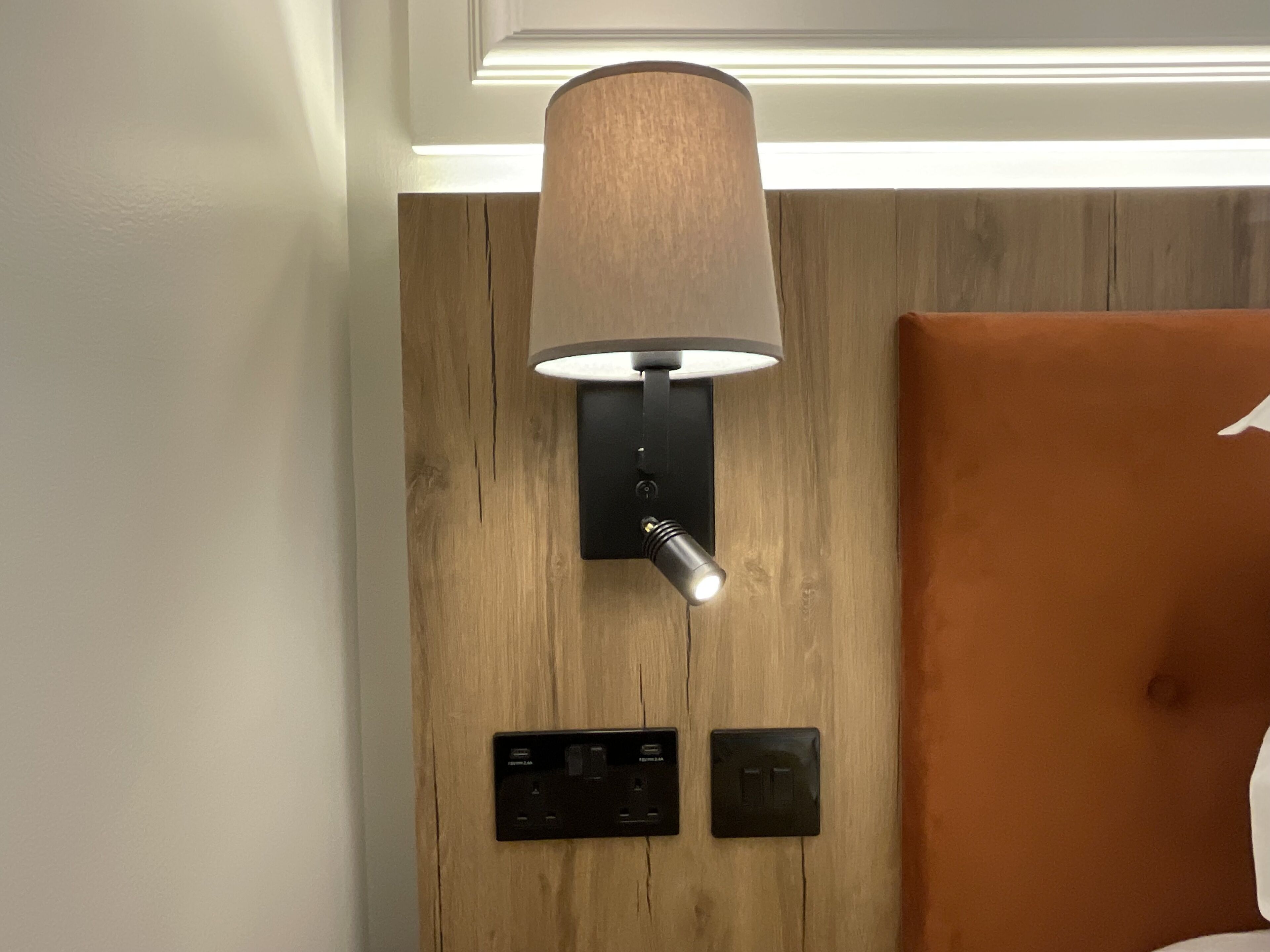 A wall-mounted lamp with a cylindrical shade, a power outlet, and a switch on a wooden panel wall.