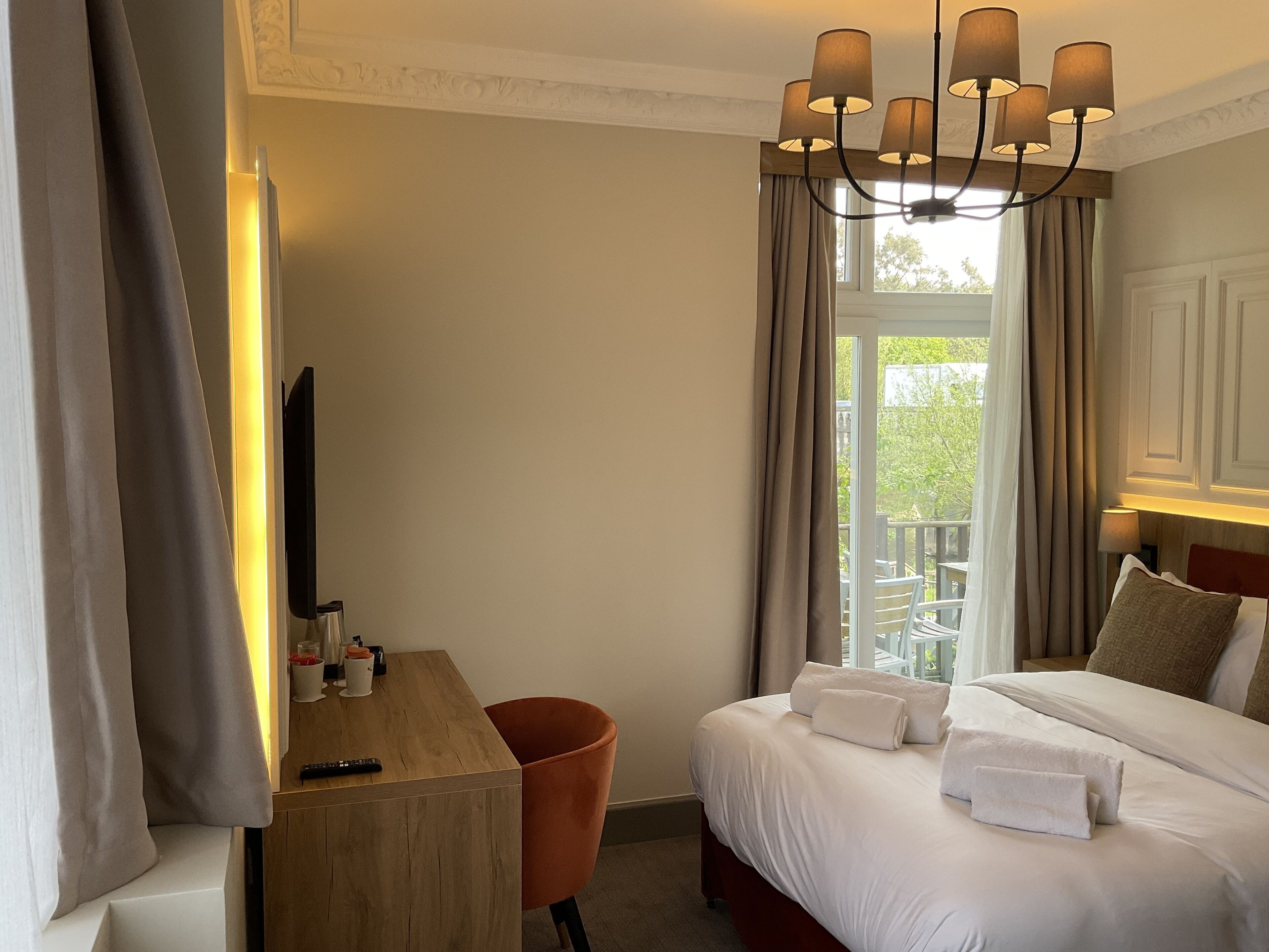 A hotel room with a bed, a desk, a chair, a TV, and a balcony with curtains.