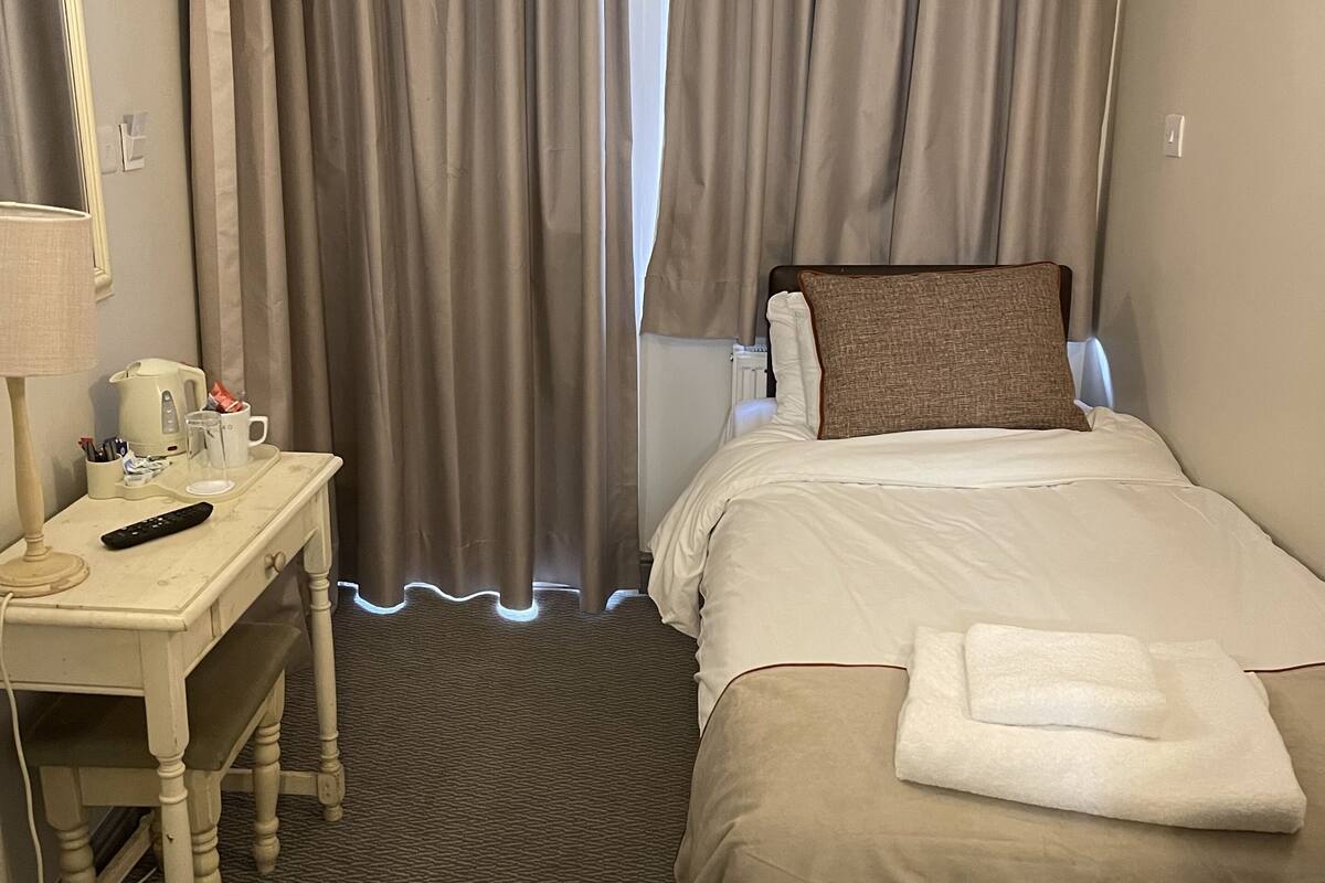 A hotel room with a bed, a desk with a lamp, a remote control, and a trash bin.