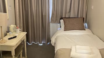 A hotel room with a bed, a desk with a lamp, a remote control, and a trash bin.
