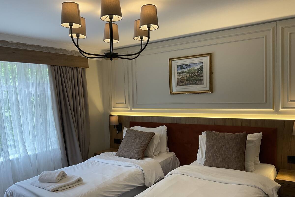 A hotel room with two beds, a chandelier, a framed picture on the wall, and a window with curtains.