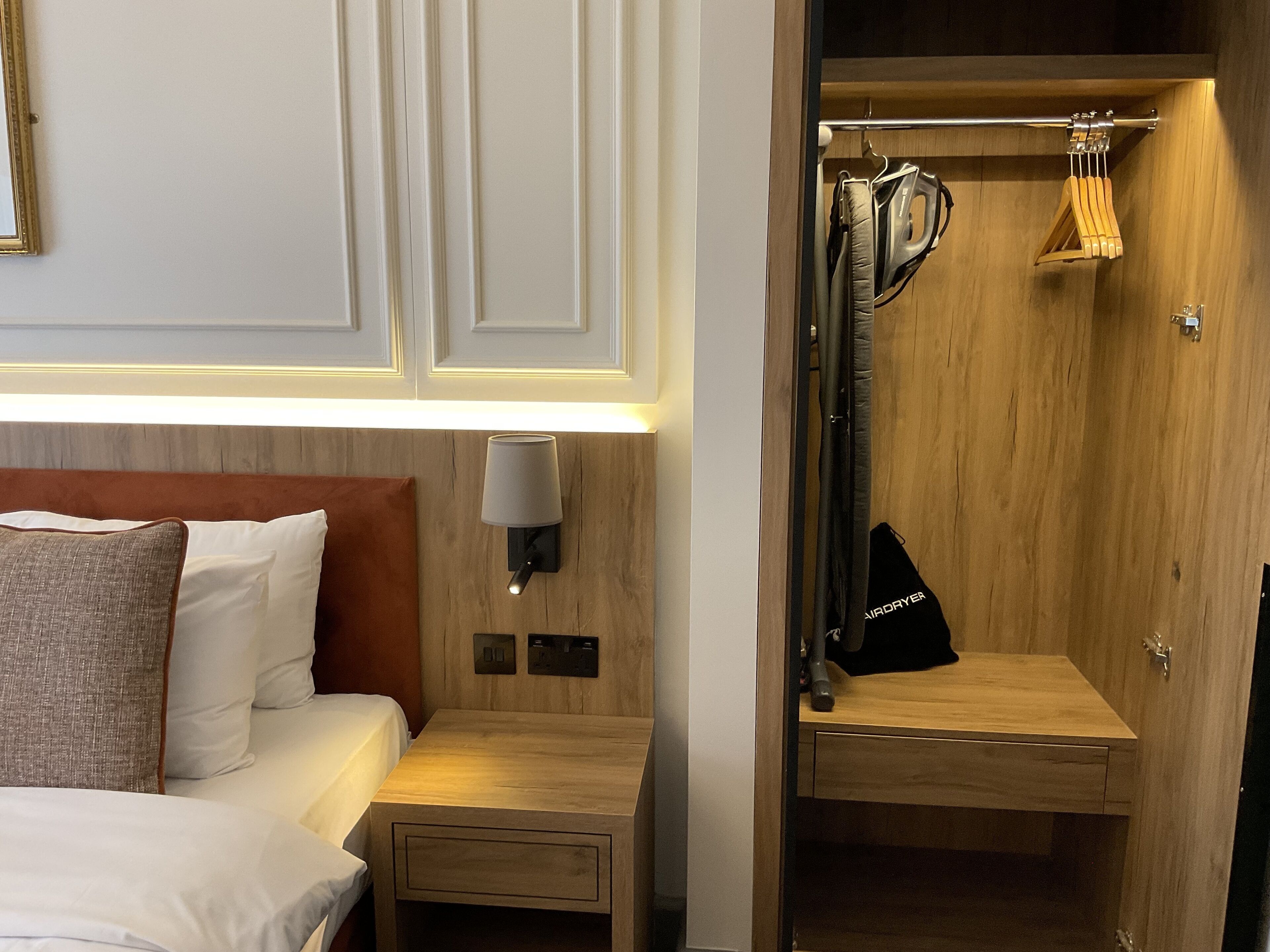 A hotel room with a bed, bedside table, lamp, and a wooden closet with hanging clothes.