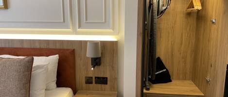A hotel room with a bed, bedside table, lamp, and a wooden closet with hanging clothes.